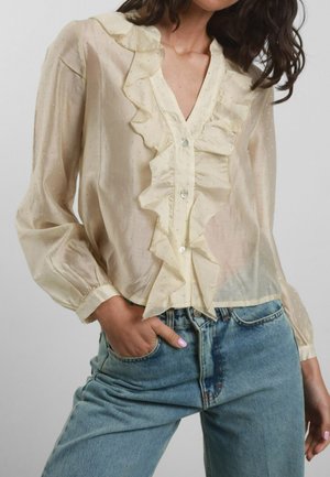 Blusa color crema con profilo arricciato sul davanti, chiusura a bottoni e maniche lunghe. Il tessuto è trasparente con sottili motivi a pois metallici. Abbinata a jeans blu.