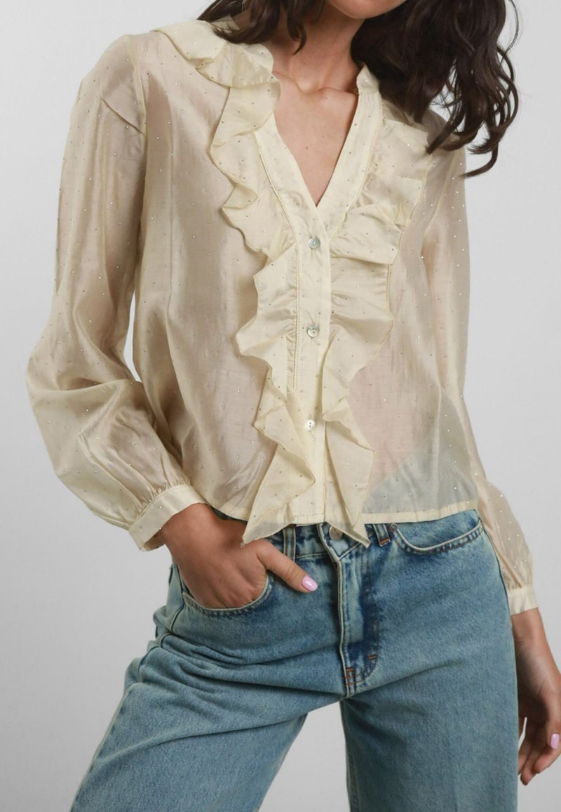 Blusa color crema con profilo arricciato sul davanti, chiusura a bottoni e maniche lunghe. Il tessuto �è trasparente con sottili motivi a pois metallici. Abbinata a jeans blu.