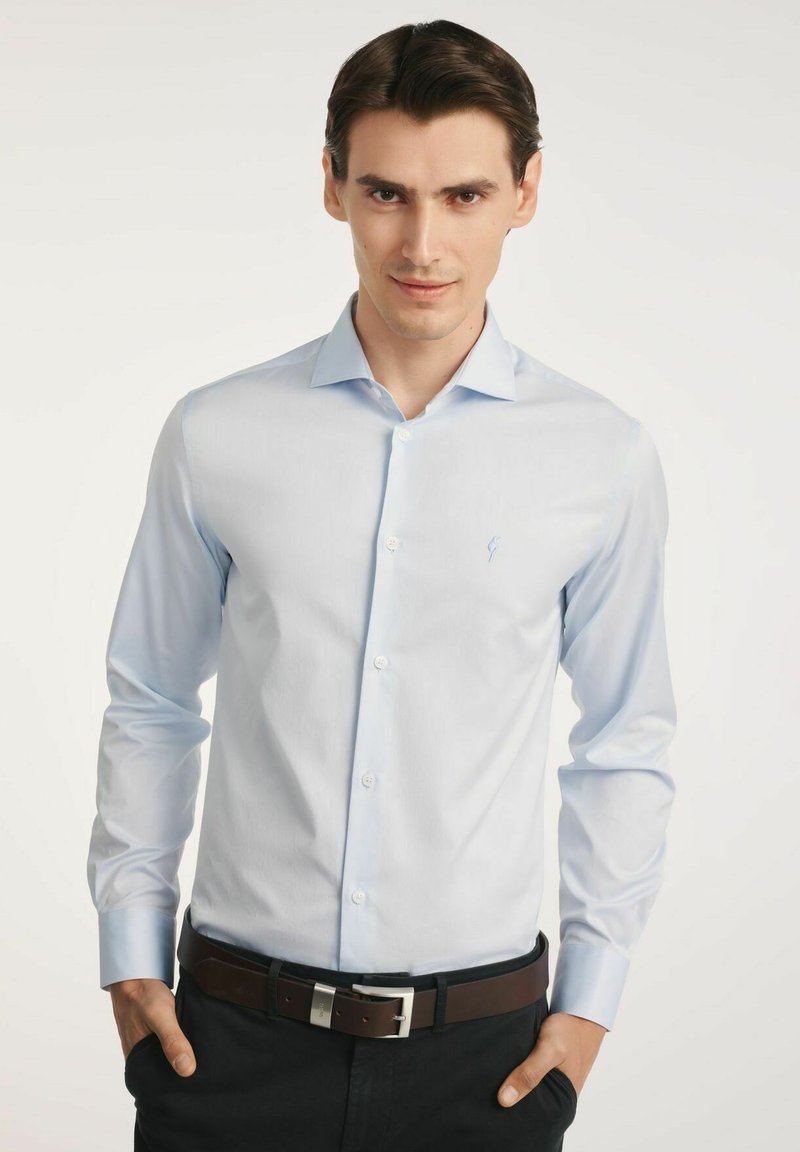 Jasnoniebieska koszula z długim rękawem, gładka w dotyku, z subtelnym logo na piersi, noszona z ciemnym paskiem i czarnymi spodniami.