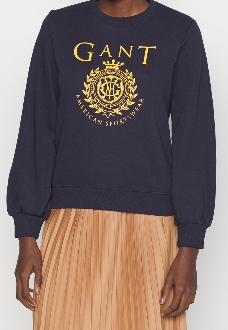 Felpa blu navy con scollatura rotonda, maniche a sbuffo e dettagli del logo stampato in oro che presenta una corona e una lauro, abbinata a una gonna in tono sabbia con pieghe.