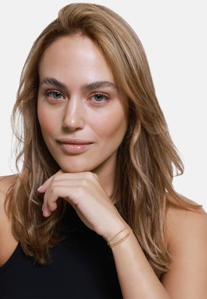 weibliches Model mit langen, gewellten blonden Haaren, das ein schwarzes Oberteil trägt. Ihre Hand stützt ihr Kinn und zeigt zwei goldene Kettenarmbänder an ihrem Handgelenk.