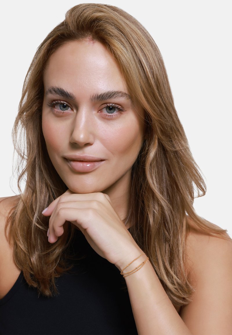 Modella femminile con lunghe e ondulate capelli biondi, che indossa un top nero. La sua mano sostiene il mento, mettendo in mostra due bracciali di catena d'oro sul polso.