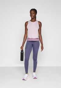 Young woman in purple leggings and light pink sleeveless top holding black water bottle, standing against plain gray background.