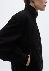 Μαύρο μπουφάν bomber από σουέτ με ψηλό γιακά, ριμπ μανσέτες και πλαϊνές τσέπες με φερμουάρ, που παρουσιάζει απαλή υφή και εφαρμοστή γραμμή.