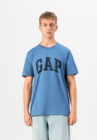 Un tânăr stă cu fața spre camera, purtând un tricou albastru cu logo-ul GAP și blugi deschiși la culoare, pe un fundal alb simplu.