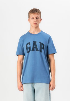 Een jonge man staat recht vooruit gekeerd in een blauw GAP-logo t-shirt en lichtblauwe jeans tegen een simpele witte achtergrond.