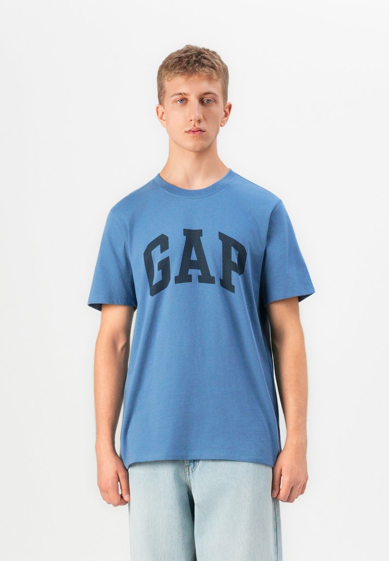 Un tânăr stă cu fața spre camera, purtând un tricou albastru cu logo-ul GAP și blugi deschiși la culoare, pe un fundal alb simplu.