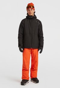 Zwarte skijas met ritszakken, gecombineerd met feloranje waterdichte broek. Zwarte handschoenen en laarzen maken de winteroutfit compleet.