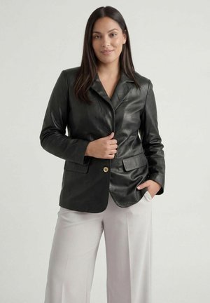 Kobieta o długich ciemnych włosach, ubrana w czarną skórzaną marynarkę i jasne, szerokie spodnie, stojąca na tle jednolitego jasnego tła.