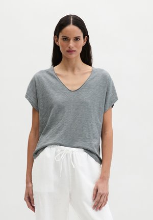 Frau mit langem dunklem Haar trägt ein graues V-Ausschnitt-Kurzarmshirt und weiße Hose mit Kordelzug, steht vor einem schlichten hellen Hintergrund.