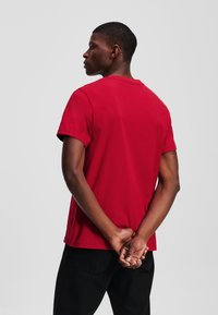 Červené tričko s krátkým rukávem vyrobené z bavlny, s kulatým výstřihem. Model stojí s crossed arms, aby ukázal design na zadní straně.