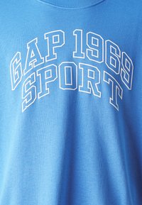 T-shirt de algodão azul claro com texto branco em destaque a dizer "GAP 1969 SPORT." Decote redondo com uma textura suave e corte descontraído.