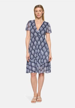 Vrouw staand, gekleed in een marineblauwe jurk tot op de knie met witte geometrische patronen en doorschijnende korte mouwen, gecombineerd met witte sandalen.