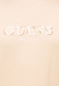 Μπεζ ύφασμα με λευκά κεντημένα γράμματα "GUESS" και από κάτω "EST. 1981", με διακοσμητική καφέ ραφή γύρω από τα γράμματα.