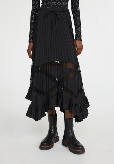 Zwarte plissérok met een rucheszoom en doorzichtige accenten, voorzien van een elastische tailleband met trekkoord. Gédragen met zwarte enkelhoge laarzen.