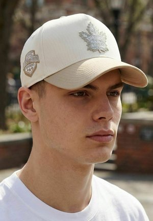 Young man wearing a beige Toronto Maple Leafs cap with a stitched logo and white shirt, outdoors with blurred background.