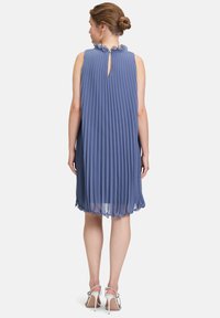 Robe plissée bleue avec un col en forme de scallop et un détail en trou de serrure au dos. Tissu transparent avec une texture douce, tombant au-dessus du genou. Talons argentés visibles.