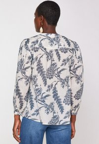 Donna vista da dietro che indossa una camicia a maniche lunghe con stampa a pappagallo blu e fiori, abbinata a jeans di denim blu.