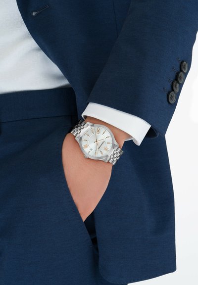 Hõbedane kell mustriga hõbedase käevõruga, valge kuupäev ja kuldsed aktsendid. Kannab kätt, mis ulatub välja sinise jakitasku.