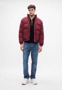Bordeaux pufferjakke med ståkrave, lynlåsclosure og et lille logo-patch, båret over en sort sweater og blå jeans.