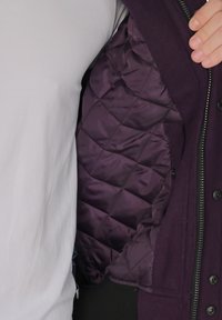 Tumma violetti eristetty takki, jossa on tikattu vuori, vetoketjukiinnitys ja nappikoristeet. Ominaisuuksiin kuuluu sileä pinta ja ommeltu kuvio sisävuorissa.