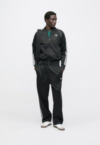 Juodas sportinis kostiumas su baltomis juostelėmis ant rankovių ir kojų. Komplekte yra užtraukiama striukė ir kelnės su elastinga juosmens juosta, derinamos su baltais sportbačiais.