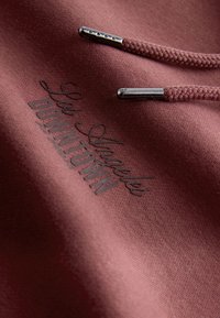 Tissu de sweat à capuche bordeaux avec cordons tressés et embouts métalliques. Présente un design en texte embossé indiquant "Los Angeles Downtown".