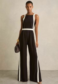 Zwarte jumpsuit met witte zijkanten, mouwloos ontwerp, brede pijpen en een getailleerde taille. Model houdt een gestructureerde zwarte clutch vast.