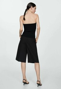 Crna rebrasta top s remenima bez naramenica uparen s crnim culottes hlačama srednje dužine, teksturirana tkanina i tanke crne sandale s potpeticama. Prikazuje minimalistički dizajn.
