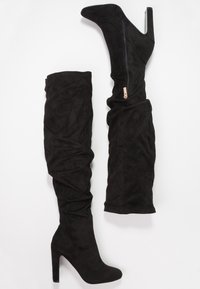 Botas negras de ante por encima de la rodilla, con puntera afilada, tacón de aguja alto y cierre de cremallera lateral con un acento dorado.