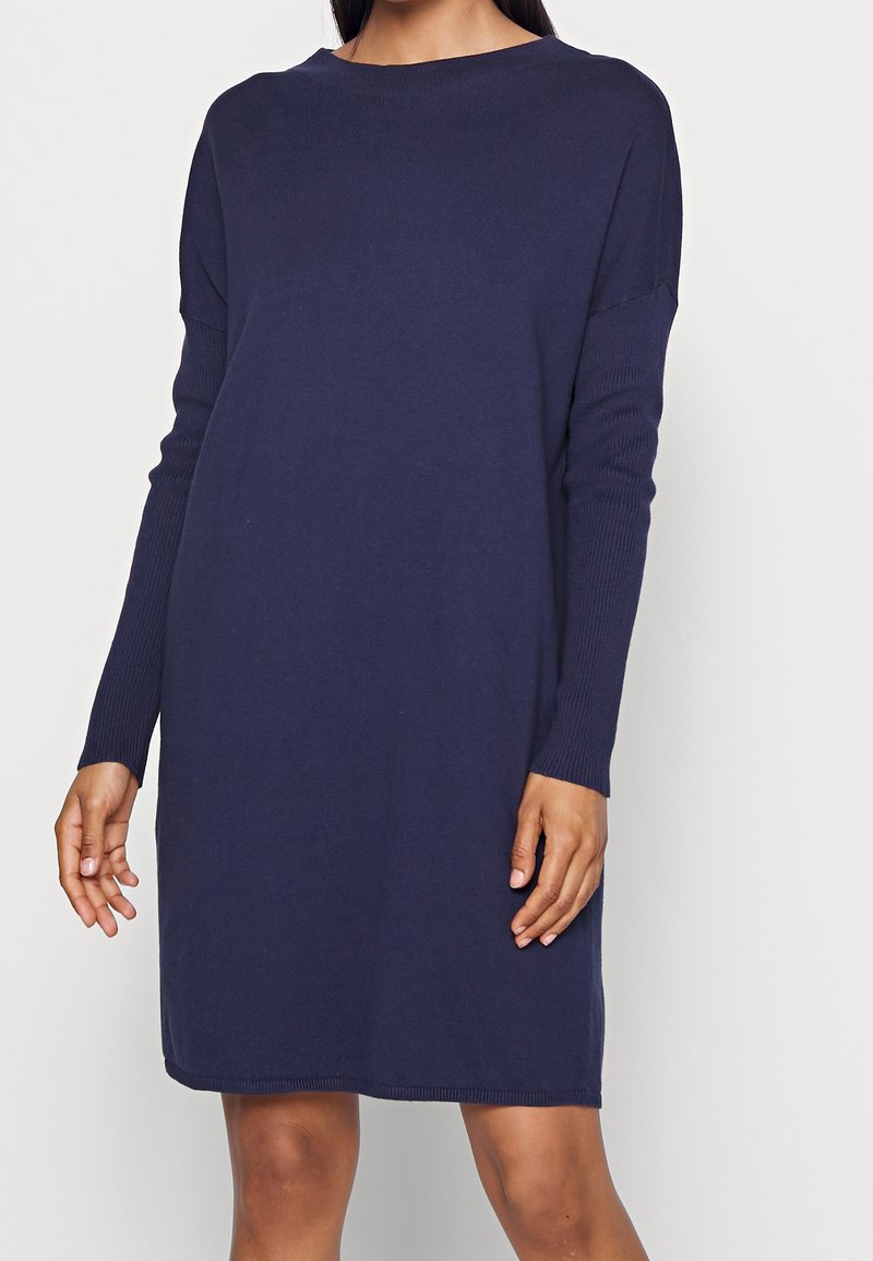 Femme portant une robe pull en maille bleu marine à manches longues avec un col bateau, debout devant un fond clair uni.