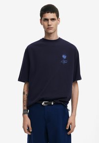 Camiseta de gran tamaño color azul marino con mangas cortas, que presenta un diseño gráfico color azul claro en el pecho. Fabricada en suave tela de algodón.