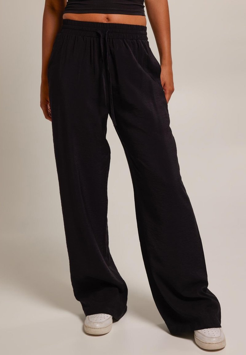 Pantalon large noir en tissu léger ; doté d'une taille élastique avec un cordon de serrage et d'une texture lisse. Des sneakers blanches sont portées.