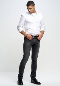 Vaqueros ajustados negros combinados con una camisa blanca de botones. El modelo lleva un cinturón negro y zapatos de vestir negros. Fondo liso.