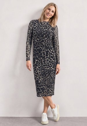 Femme aux cheveux blonds, portant une robe midi à manches longues imprimée léopard et des baskets blanches, debout contre un mur clair.