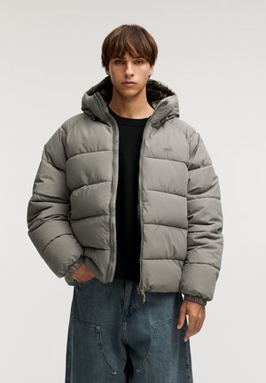 Gray padded jacket with a hood, zip closure, elastic cuffs, and two side pockets. Worn over a black sweater and loose denim pants.