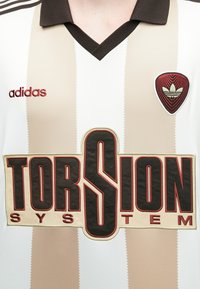 Πουκάμισο με λευκές και μπεζ ρίγες και V-λαιμόκοψη, με σκούρο γιακά. Λογότυπο "TORSION SYSTEM" σε κόκκινο και μαύρο, με έμβλημα Adidas από πάνω.