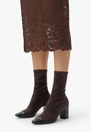 Braune Wildleder-Stiefeletten mit schwarzen Kappen, die einen enganliegenden Schaft und einen klobigen schwarzen Absatz aufweisen. Kombiniert mit einem braunen Spitzennrock.