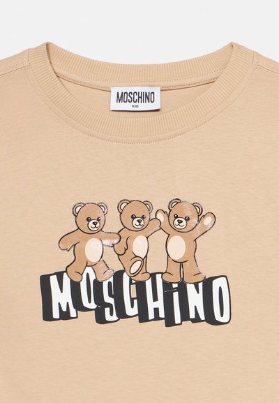 Μπεζ παιδικό μπλουζάκι με τρία αρκουδάκια που χορεύουν πάνω από το ασπρόμαυρο κείμενο "MOSCHINO" στον μπροστινό λαιμό.