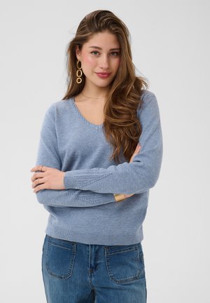Jeune femme aux longs cheveux bruns portant un pull bleu clair, un jean bleu, des boucles d’oreilles en or et un bracelet, debout les bras croisés.