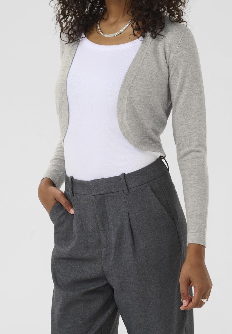 Femme portant un haut blanc sans manches, un cardigan court gris clair, un pantalon taille haute gris foncé et un collier chaîne argenté.