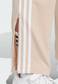 Beige träningsbyxor med vita sidostråk, med en dragkedja vid vristöppningen som avslöjar en svart- och vitrandig strumpa. Vit sneaker nedanför.