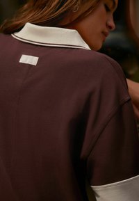 Braunes Poloshirt mit einem weißen Kragen. Der Stoff wirkt weich und hat eine glatte Textur. Hat ein kleines Etikett auf der Rückseite.