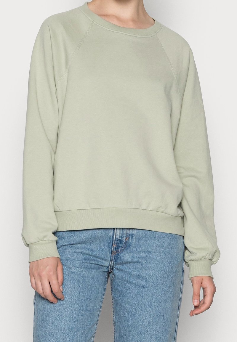 Felpa verde chiaro con colletto a girocollo a coste e polsini, orlo arrotondato e maniche a raglan, abbinata a jeans azzurri.