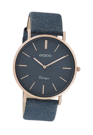 Montre ronde avec un cadran texturé bleu foncé, des aiguilles et des index en or rose, et un bracelet en cuir bleu doux. Présente le texte "Vintage" au-dessus du centre.