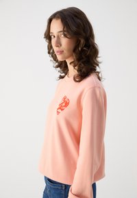 Rosa hettegenser med lange ermer og rund hals, med et trykt hjerte design i rødt på venstre brystområde.