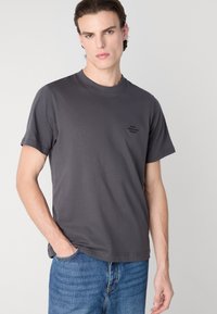 Grå bomull t-skjorte med korte ermer, rund halslinning og en liten svart logo på venstre bryst. Kombinert med lyseblå jeans.