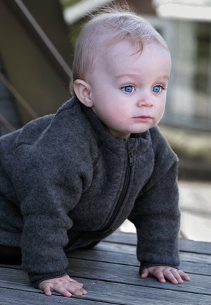 Veste en polaire grise avec col montant et fermeture éclair, portée par un enfant qui rampe sur une surface en bois. Les caractéristiques notables incluent un matériau texturé et des manches avec poignets.