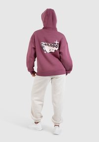 Felpa con cappuccio viola che presenta un grande design stampato sul retro, abbinata a pantaloni jogger grigi chiari e sneakers bianche.