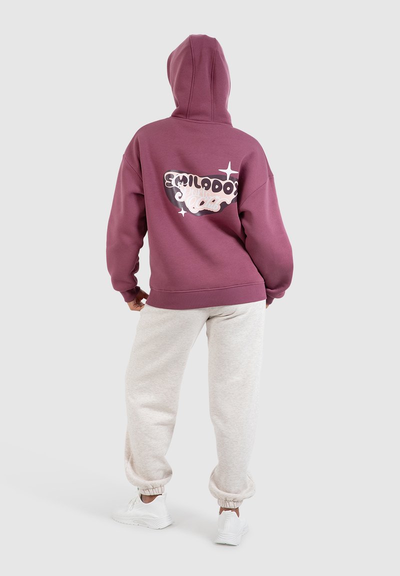 Lila Kapuzenpullover mit einem großen Druckdesign auf der Rückseite, kombiniert mit hellgrauen Jogginghosen und weißen Turnschuhen.
